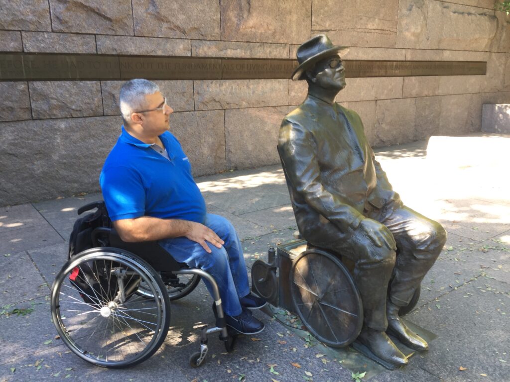 Fadi, outdoors and seated In his wheelchair, regards a life-size statue depicting President Franklin D. Roosevelt also seated in a wheelchair. Fadi wears a blue polo shirt and jeans.