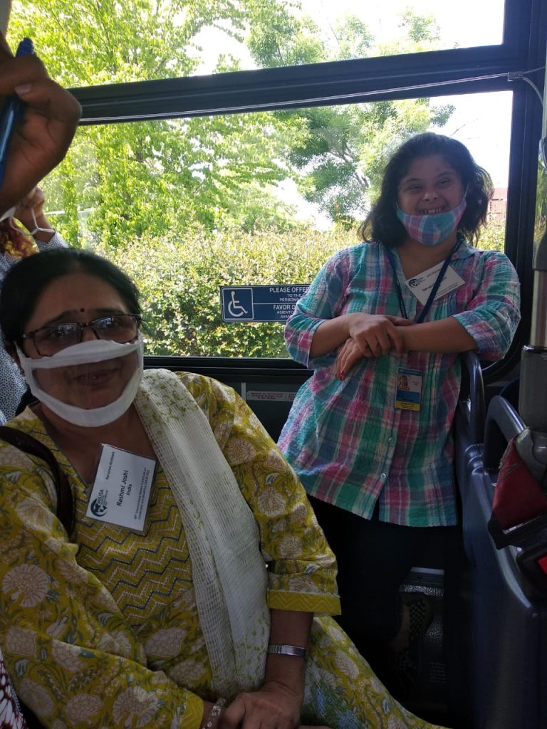 Two women from India smile while on a public bus. Both wear nametags and facemasks. 