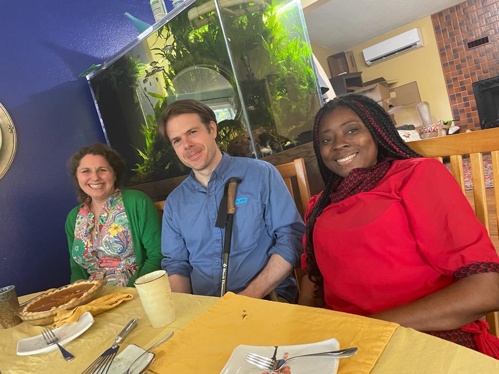 Three people, including one woman from Cameroon and a man and woman from the U.S., sit at a table inside a home. On the table is a pie and serving utensils. Behind them is a small aquarium with several fish swimming.