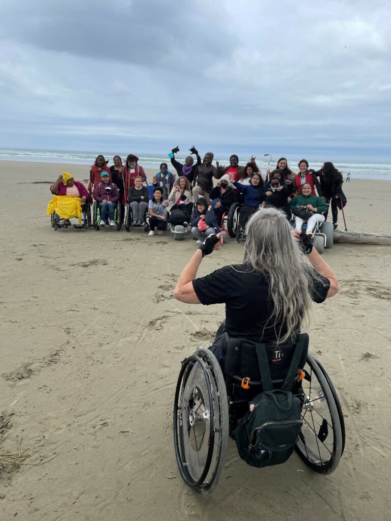 A woman riding a wheelchair faces a group of 22 women smiling, all who are WILD delegates. She hold her fists up. Some women have their arms up, some are riding wheelchairs, some are holding canes. They are at the beach on the sand with the ocean in the background.