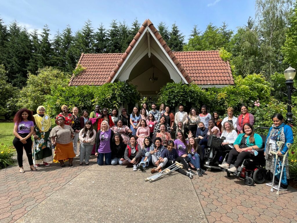 WILD delegates, GDDI attendees, and MIUSA staff pose together smiling while outdoors in a courtyard surrounded by trees. Some women use wheelchairs and power scooters, and some women hold crutches.