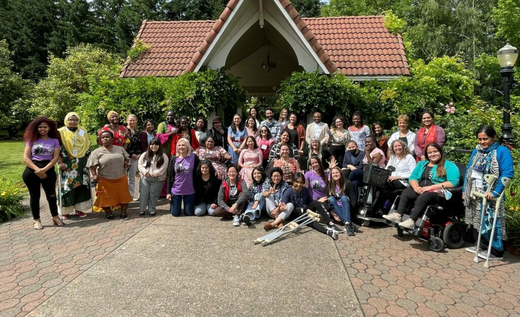 WILD delegates, GDDI attendees, and MIUSA staff pose together smiling while outdoors in a courtyard surrounded by trees. Some women use wheelchairs and power scooters, and some women hold crutches.