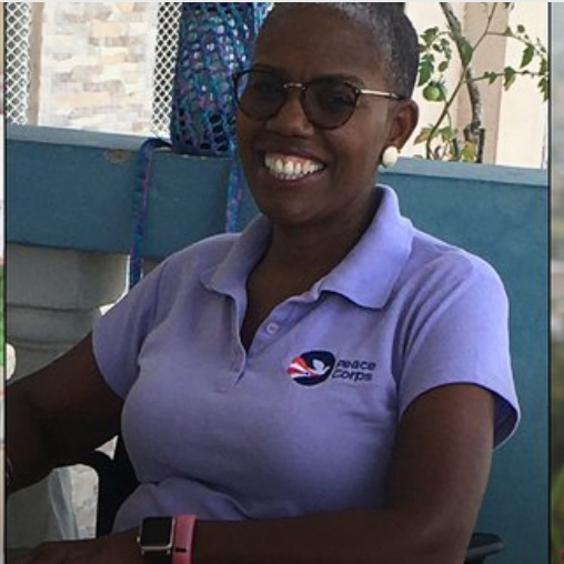 Judy, a black woman with glasses and black buzz cut and a white earring smiling at the camera. She wears a light lavender polo shirt.