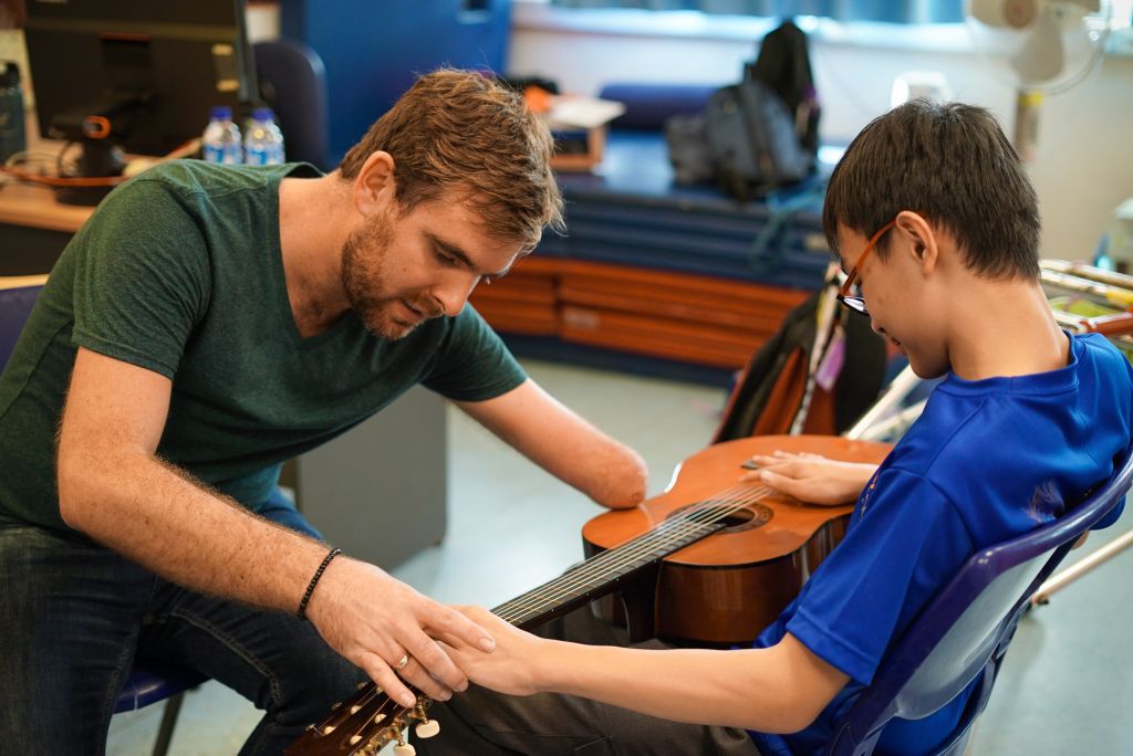 Tony a white man with brown hair and a beard, wears a green shirt as he is instructing a young boy on where to place his hand to play the guitar on the young boy's lap. Tony has one short arm missing a hand placed on top of the body of the guitar.