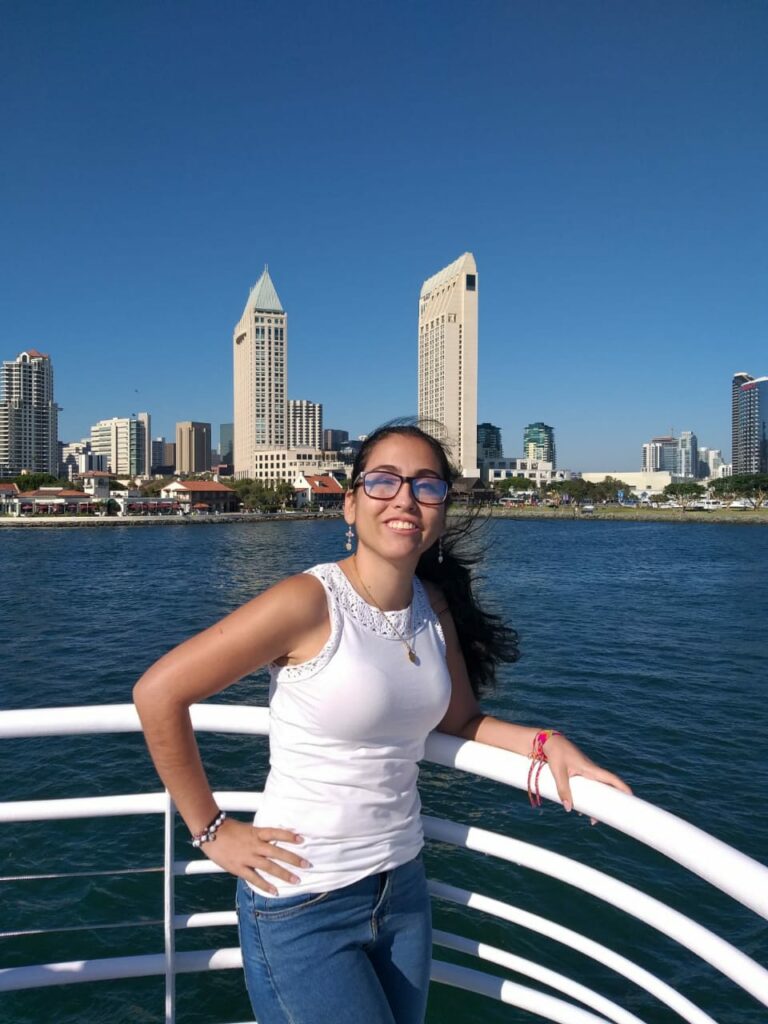 Sara, a Latina woman with long black hair in a ponytail and glasses smiling at the camera; she wears a white short sleeve blouse and dangling earrings