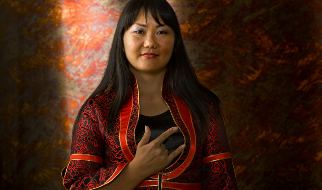 Professional color portrait of young East Asian woman in red and black gown, smiling serenely at the camera, holding "I Love You" sign language sign hand over her heart