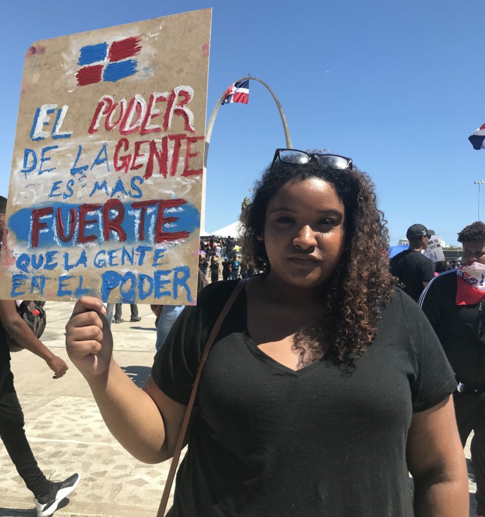 Doris wears a black shirt, and her glasses on her head. She wears a serious look and holds a cardboard sign with hand-painted red, white and blue letters with the phrase “El poder de la gente es mas fuerte que la gente en el poder”.