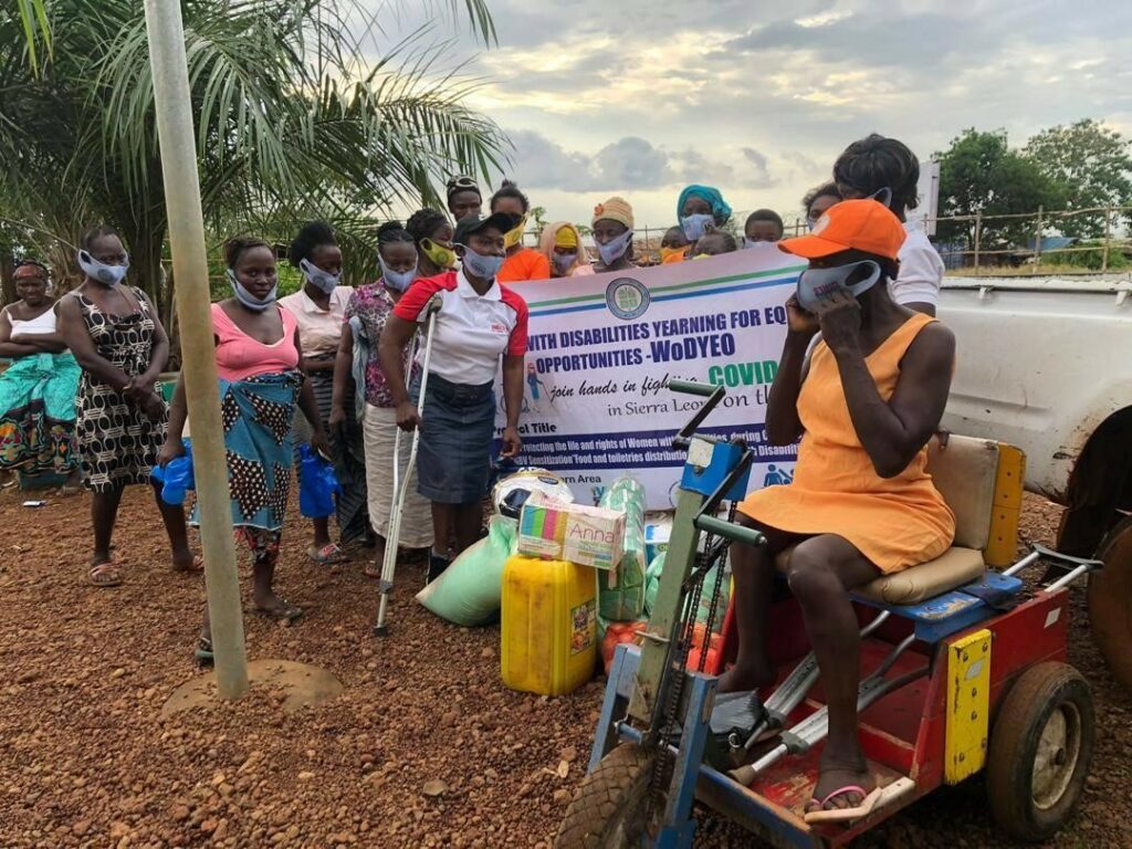 A group of young and middle aged women, some with visible disabilities, gather around a banner whose words are partially obscured, near a mound of equipment and gear.