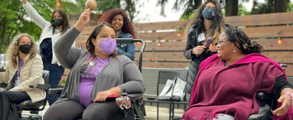 Women gather on a dance floor among a wooded setting. In foreground, two young adult women with physical disabilities share a joyful moment. Some women shake percussive musical instruments.