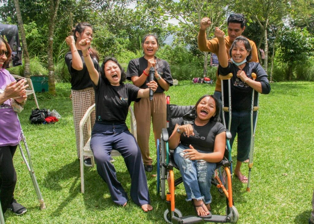 Five women participants of WILD-Indonesia sit and stand together holding their fists in the air, smiling and shouting. One participant is sitting in a manual wheelchair and one participant is standing with crutches.
