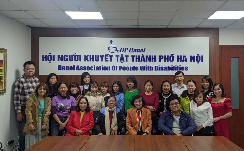 Participants of WILD-Vietnam smile for a group photo in a conference room. Behind the group is a banner that shows an image of an accessibility logo and the words "DP Hanoi, Hanoi Association of People with Disabilities."