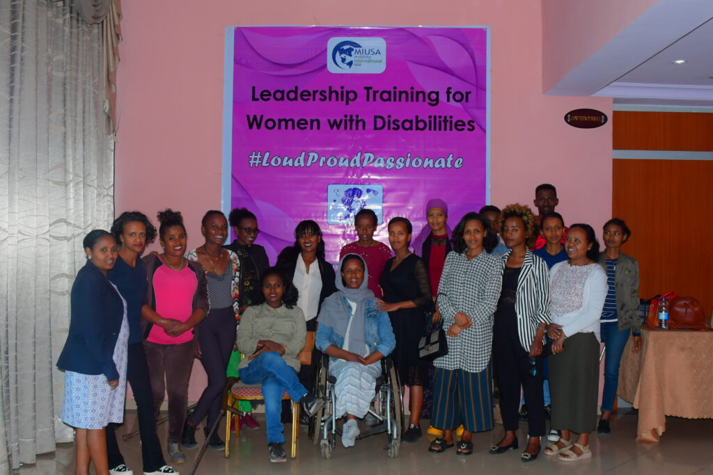 Twenty women with diverse disabilities smile facing the camera in a conference room. Behind them is a bright pink and purple poster with the MIUSA logo, EDWNA logo, and the words, "Leadership Training for Women with Disabilities #LoudProudPassionate"