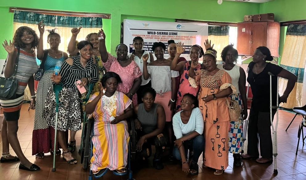 A group of women with various disabilities sit and stand in a room facing the camera, smiling. Some are using crutches and one woman is using a wheelchair. Several women hold their arms up waving.