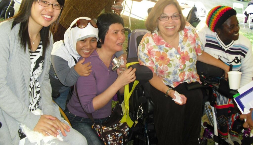 Judy smiles at camera as she sits with a group of four women with apparent and non-apparent disabilities from different nationalities, races/ethnicities.