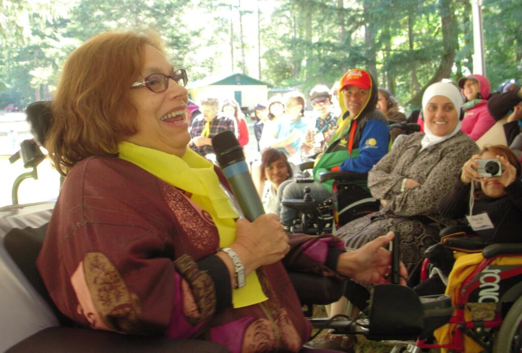 Judy laughs, holding a microphone, surrounded by a crowd of women, several seated in wheelchairs. They are under a large unseen canopy tent with tall green trees in background.