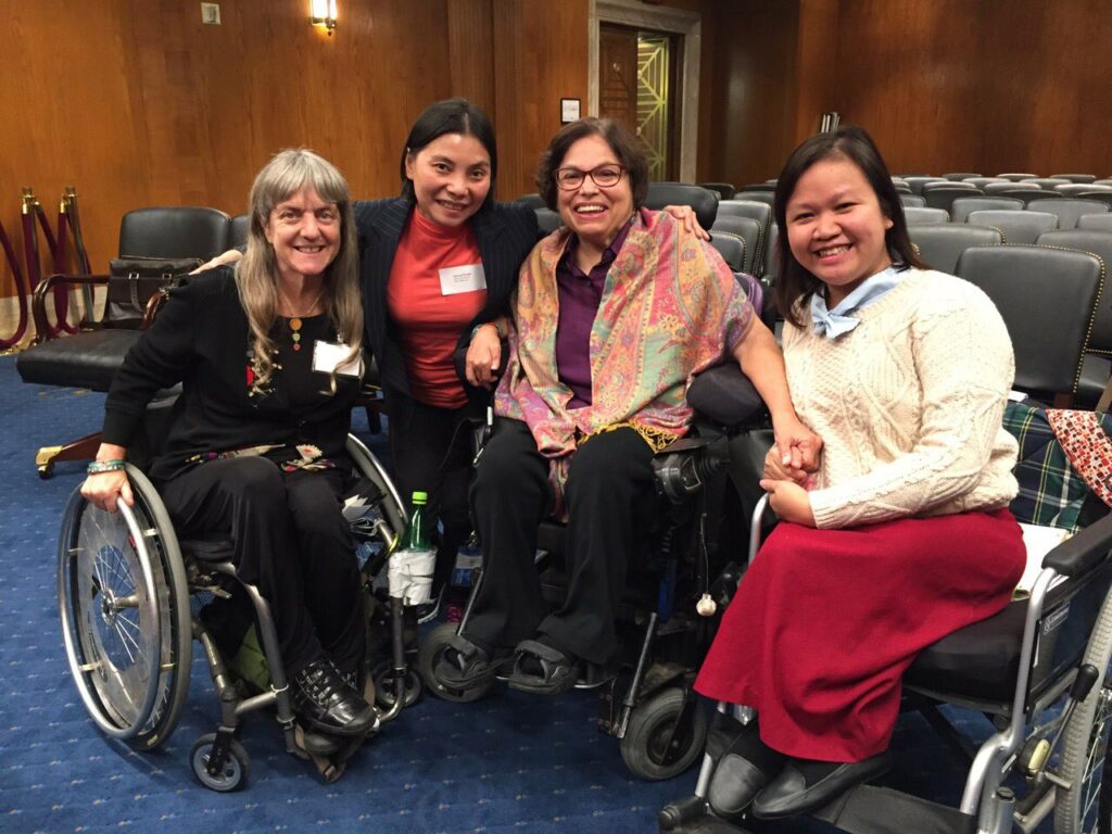 Four women seated next to one another in an otherwise empty conference room, smiling. Three women use wheelchairs.