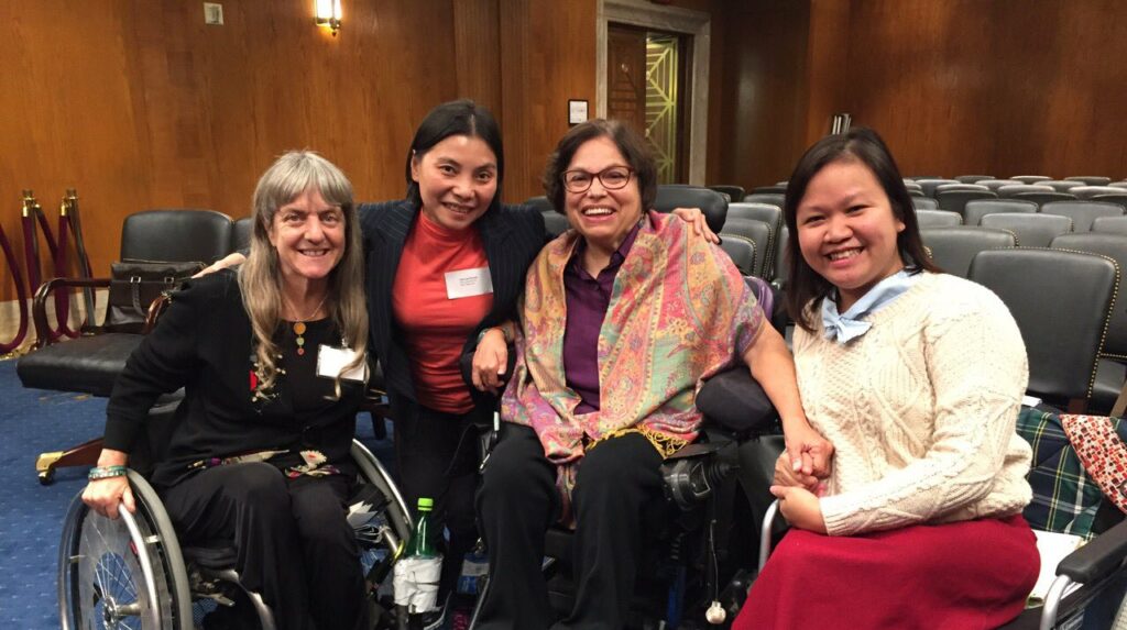 Four women shoulder to shoulder in conference room, from left to right: Susan Sygall, a WILD alum from Vietnam, Judy Heumann, another WILD alum from Vietnam