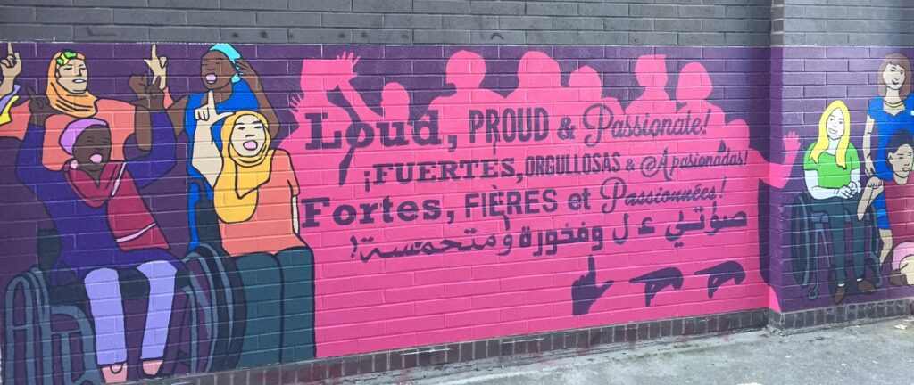 A colorful mural on a brick wall shows a group of women with different disabilities and diverse backgrounds singing and signing. The words "Loud, Proud and Passionate!" appear in English, Spanish, French and Arabic.