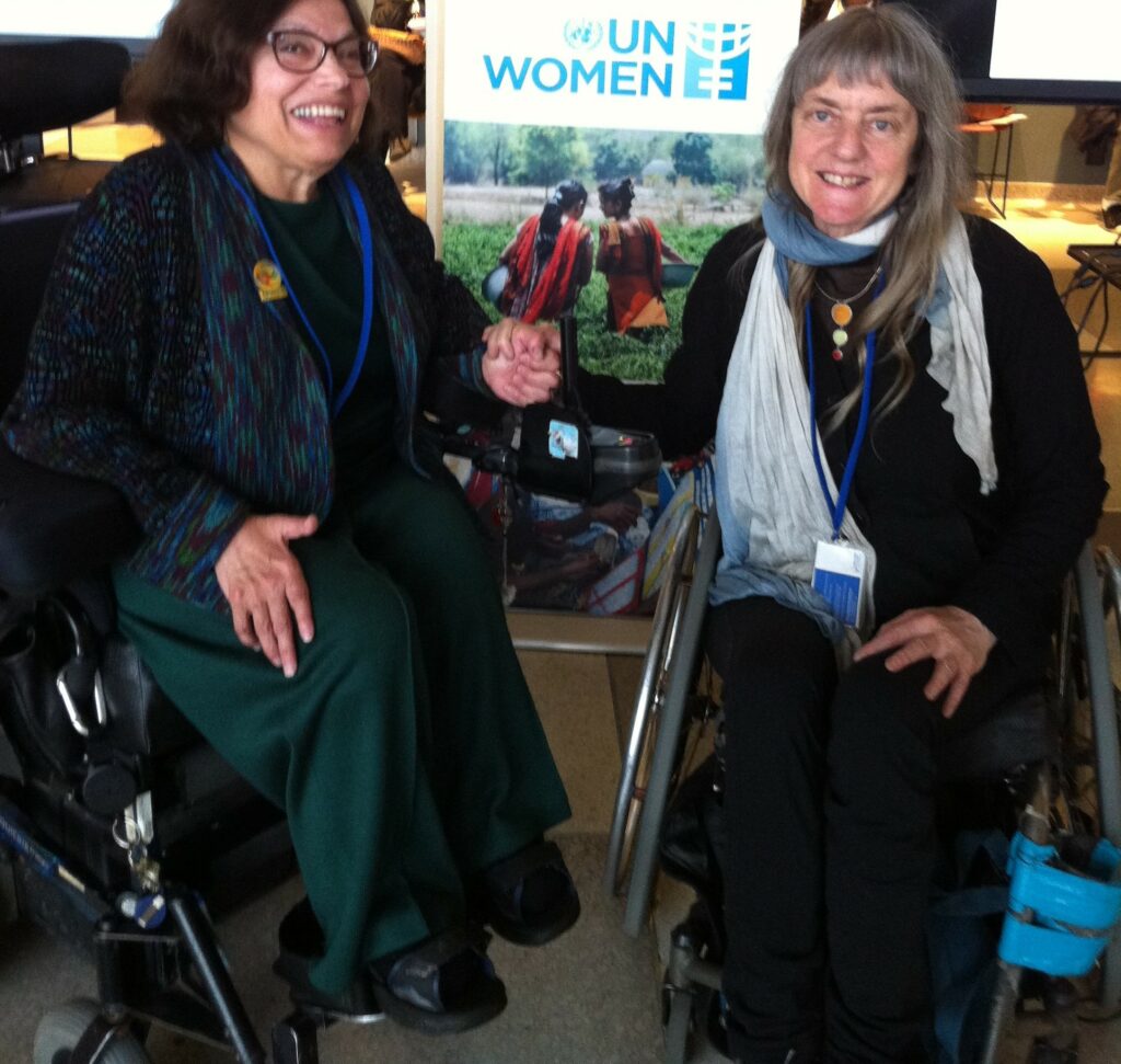 Judy and Susan Sygall clasp hands in front of a banner that reads "UN Women". Both wear semi-formal attire and are seated in wheelchairs