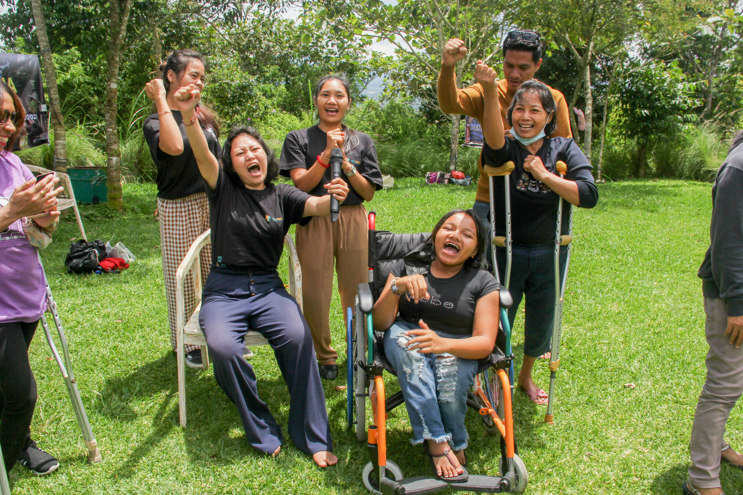 Five women with various disabilities sit or stand together holding their fist in the air while shouting and smiling. They are in an outdoor park, on green grass with trees in the background.