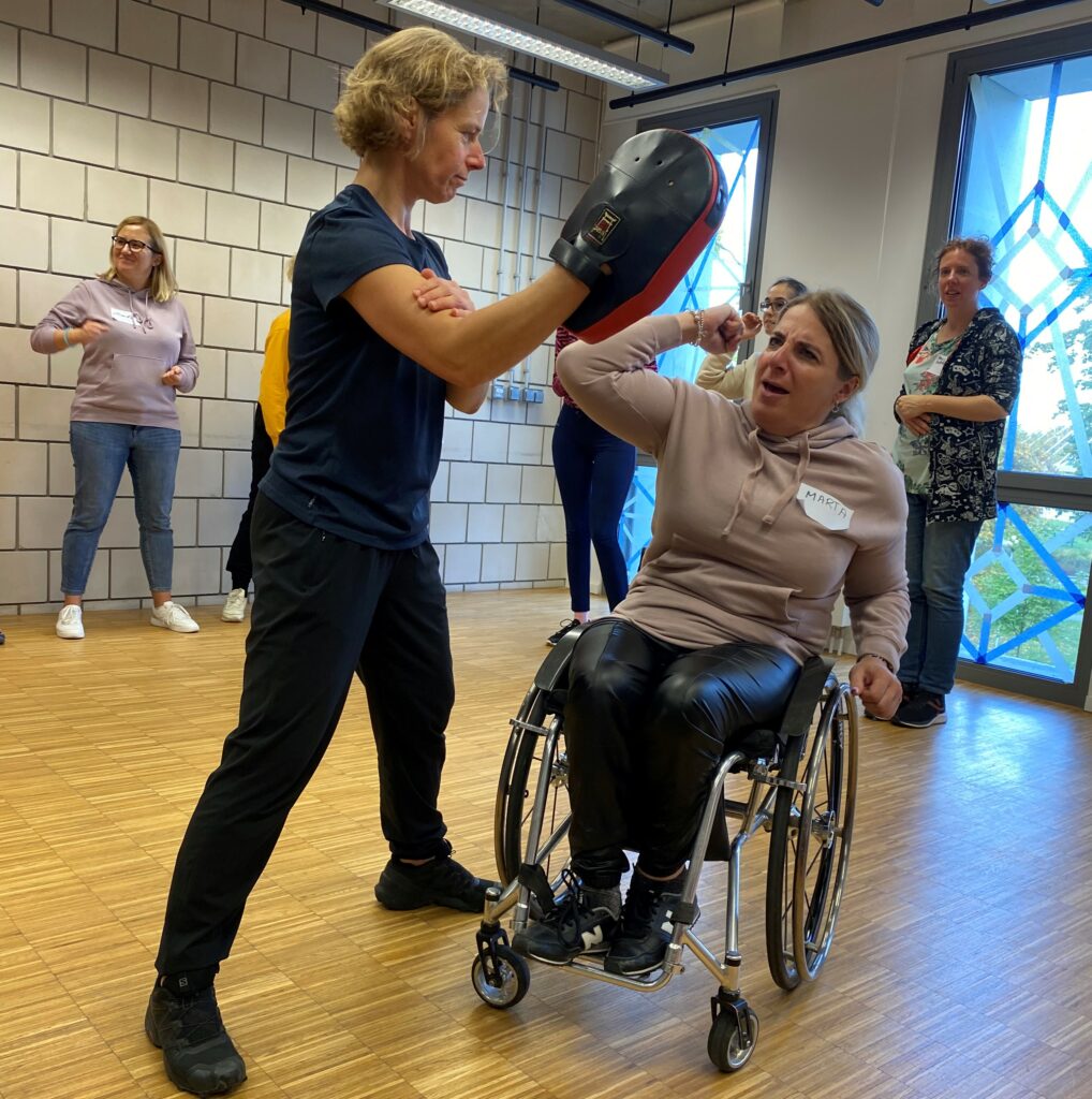 Woman using a manual wheelchair raising her arm with her elbow bent and her hand in a fist. Another woman standing in front of her holds a padded cushion.