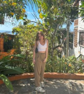 White woman with long, light brown hair wearing tan pants and a white tank top standing in a courtyard with tropical, flowering trees and houses on the edge and the ocean in the distance.