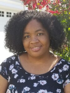 Black woman with short, curly hair wearing a blue blouse with white flowers smiling.  A ref flowing plant is behind her.