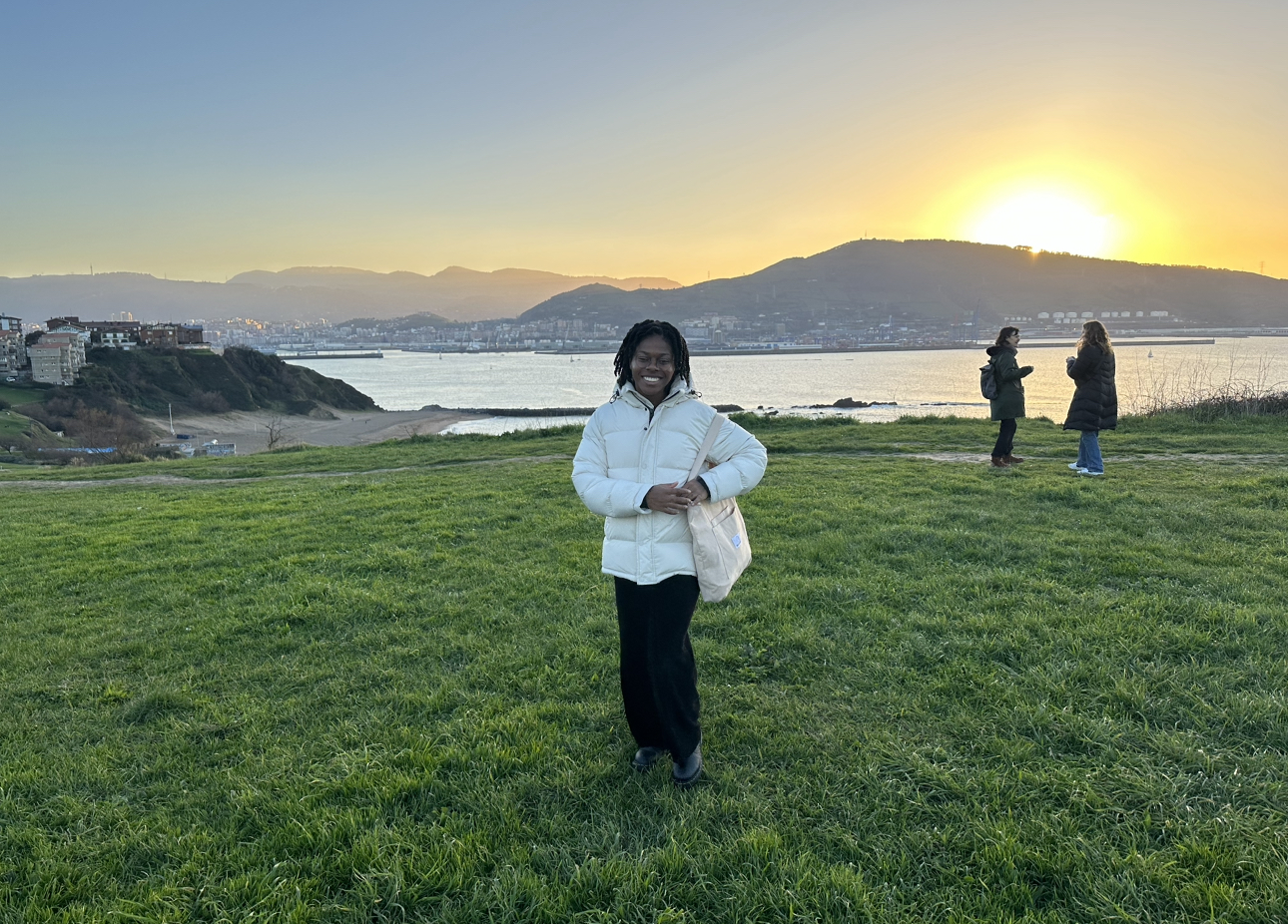 Black woman wearing a white jacket and black pants standing in a green field with the ocean bay in the background, a hill behind the bay and the sunsetting. Two people stand facing each other behind Abena. 