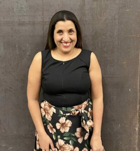 Moroccan woman with shoulder-length black hair smiling and wearing a black top and flowered skirt. 