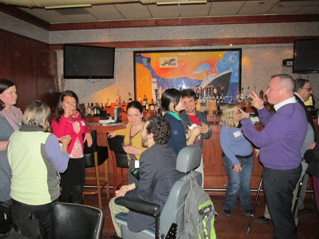 A group of people, some seated and some standing, gathered in a room talking to one another. One person uses a power wheelchair.