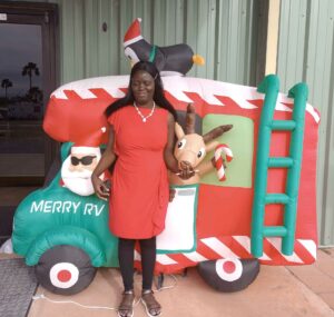 Black woman wearing red dress standing in front of an infalted red, green, and white truck with Santa Clause driving and the words movin on below the driver's window. A penguin with a Sant's hat is on the top. Kadije is holding an inflatable reindeer head under her right arm.