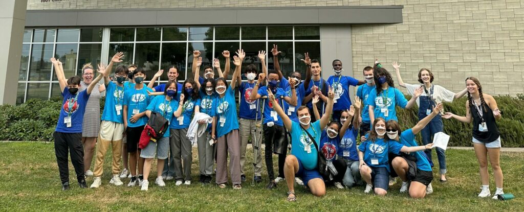 Group of high school exchange students pose in front of a high school building. They are smiling and waving their arms.