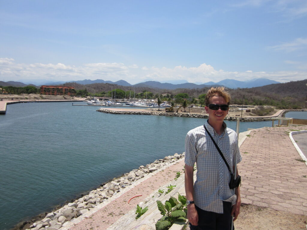 White male with blond hair blowing in the wind wearing dark glasses, holding white cane smiling in front of marina standing on stone and sand pathway.