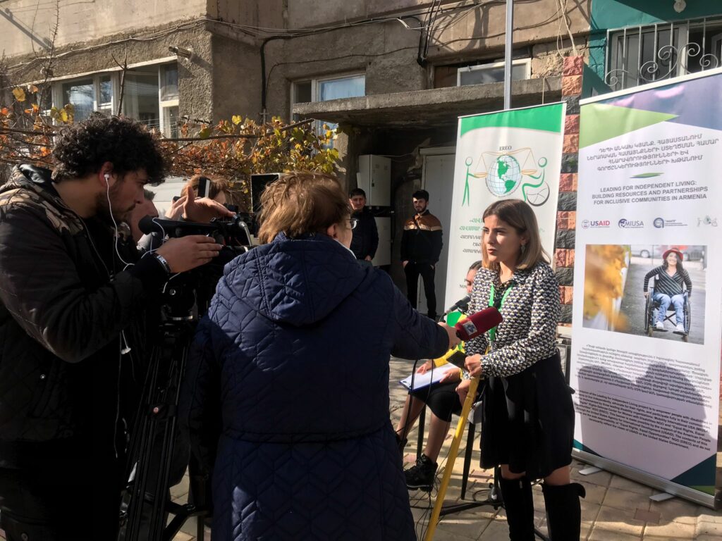 Anush Aslanyan stands outside of the ILRC building talking to three journalists. Two journalists hold cameras and one holds a microphone.