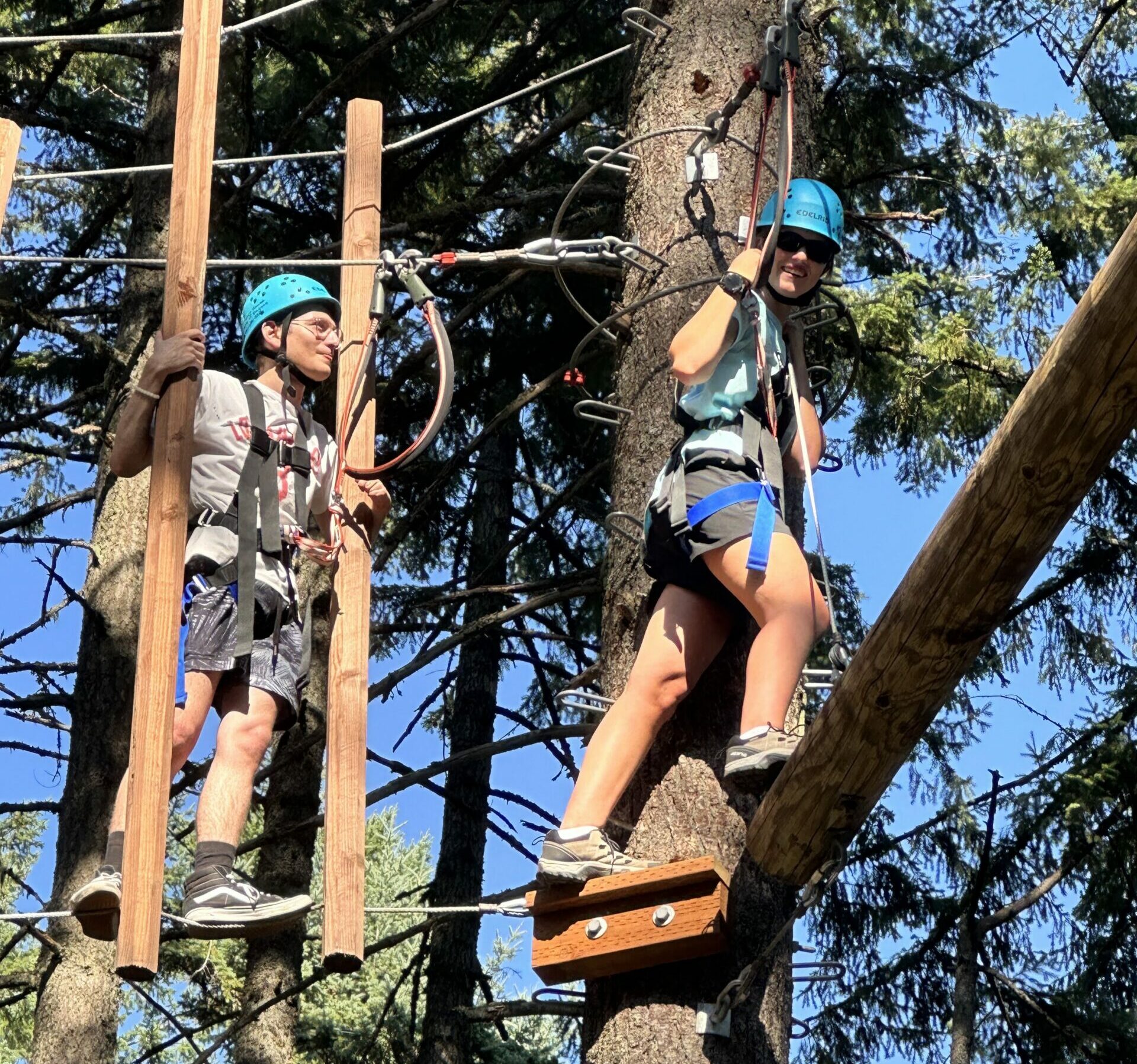 Two students wearing harnesses and helmets holding onto ropes and wooden columns while climbing between trees on the challenge course. A tall tree is between them.