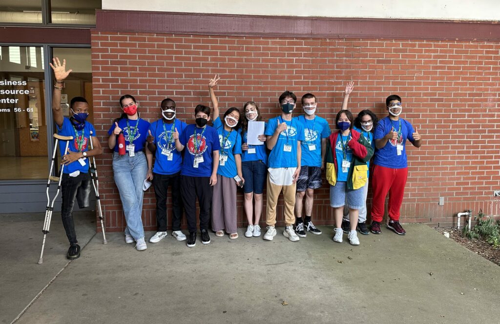High school exchange students stand side by side against a brick wall, waving or smiling at the camera. They wear COVID prevention masks and one student uses crutches