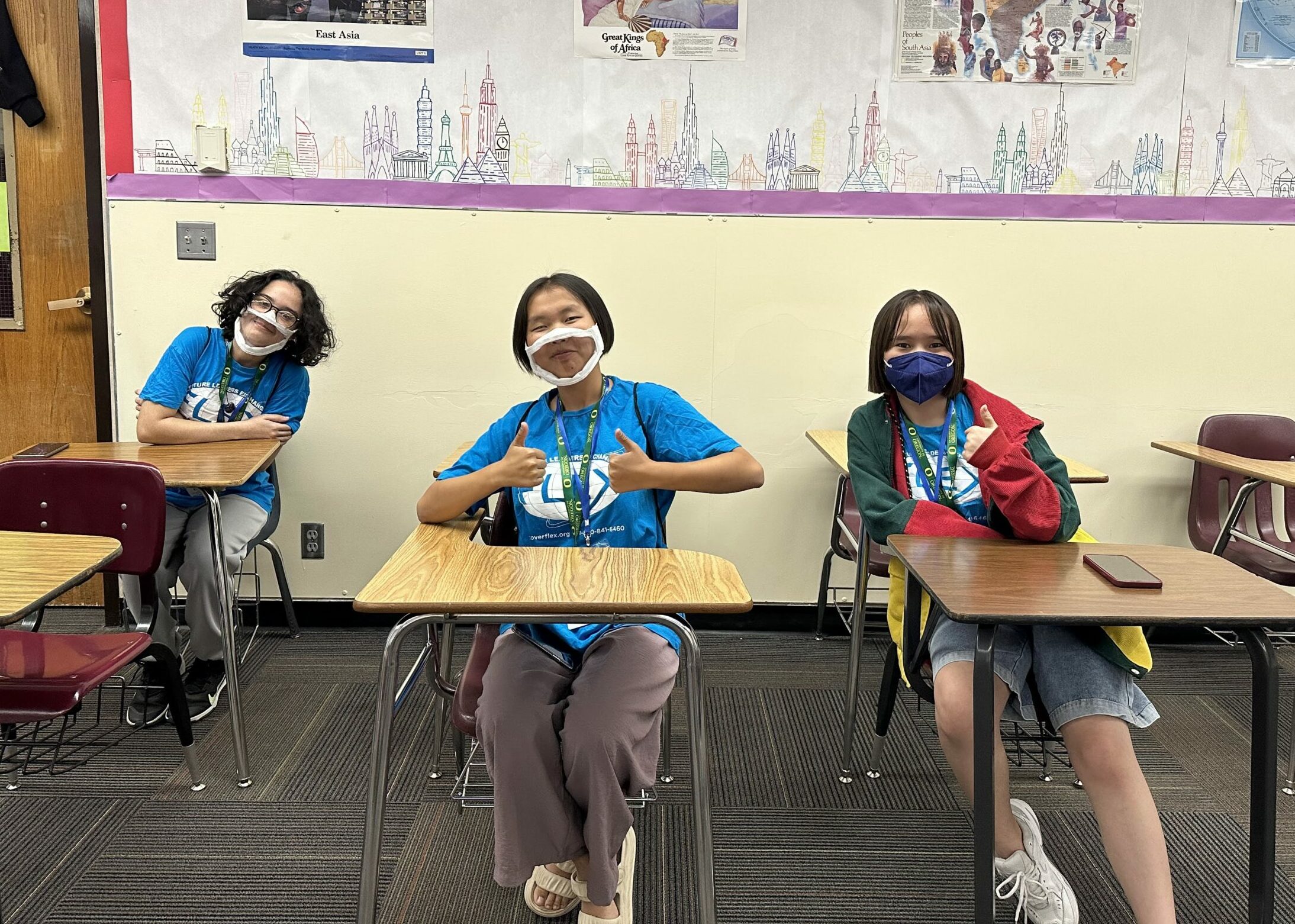 Three students sit at desks inside a classroom. They are smiling towards the camera. One student holds two thumbs up.