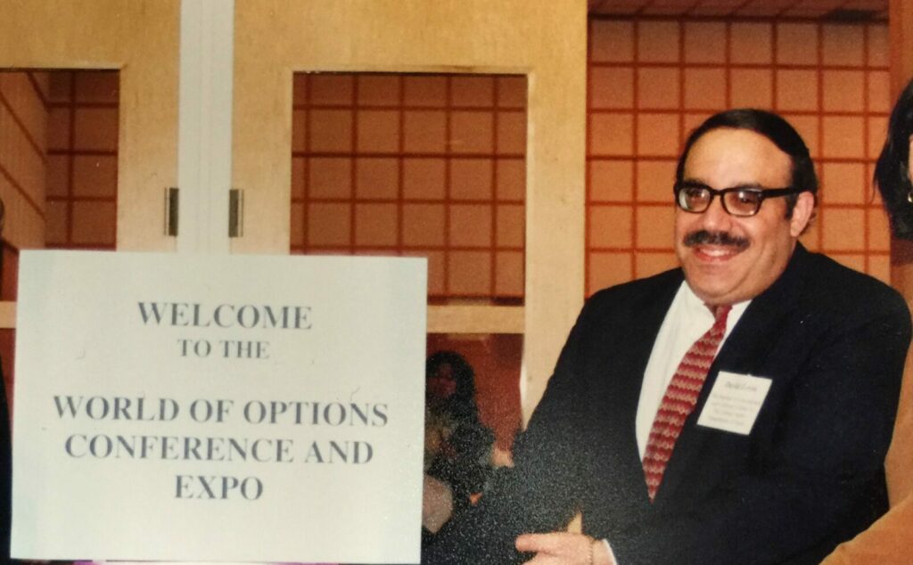 Vintage photo of David Levin in perhaps his 40s, wearing suit and tie and moustache, standing indoors pointing to a sign that says Welcome to a World of Options Conference