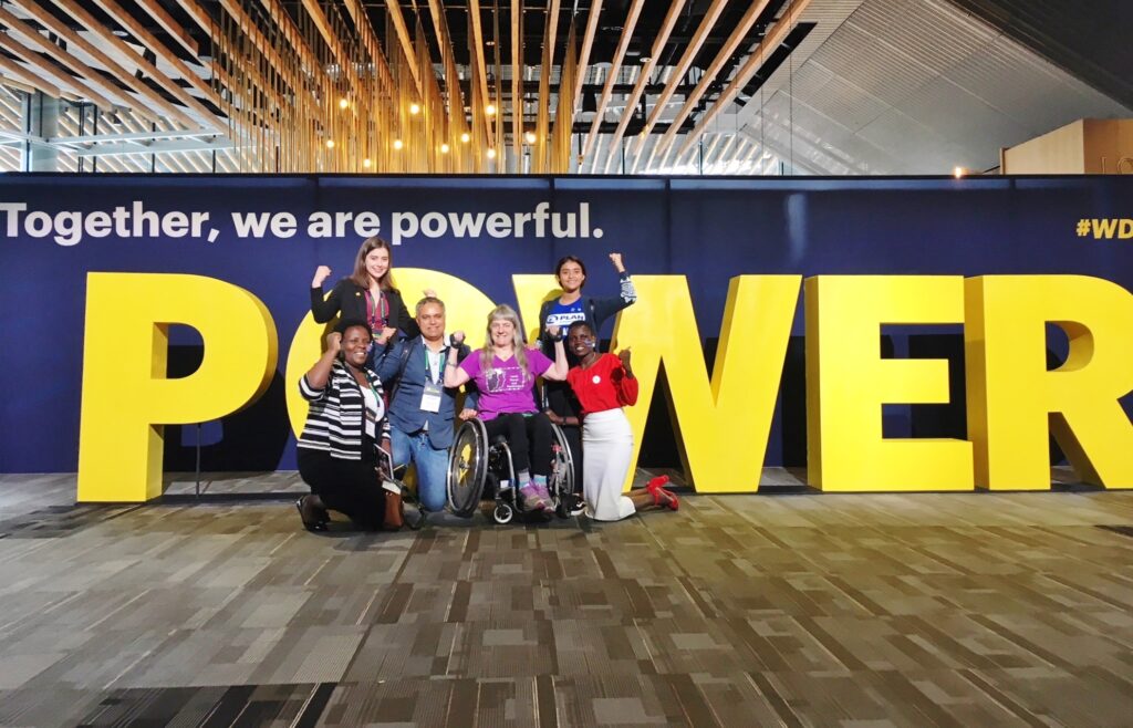 Susan Sygall with a group of 5 people gathered in front of a large yellow "POWER" sculpture/sign. Susan and the others hold up their arms in powerful flexing motion.